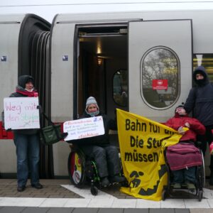 Protestaktion u.a mit Banner "die Stufen müssen weg" vor der Tür eines ICE, zwei Menschen im Rollstuhl dabei zwei Schaffner*innen gucken in die Richtung der Aktion