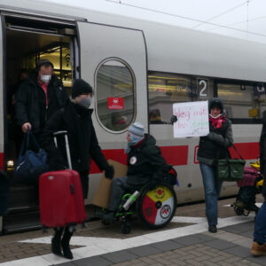 Rollstuhlfahrerin an der Tür eines ICE mit Stufen, Menschen steigen aus. Im hintergrund eine Person mit Plakat " Wer mit den Stufen"
