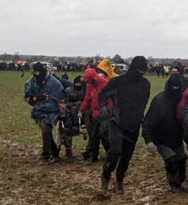 menschen tragen eine Person im Rollstuhl auf einem Acker, in Hintergrund andere Demonstrierende