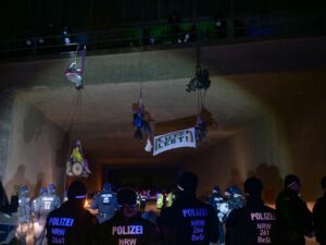 menschen hängen in seilen an einer Brücke, zwei sind im Rollstuhl im Seil, es gibt viel Polizei. Ein Banner Lützi Lebt
