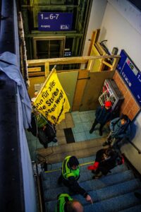 eine Person hängt im Rollstuhl sitzend in Seilen an einer Bahnunterführung, hinter ihr eine Aufzugbaustelle, davor die Treppe zum Gleis und DB Siherheit. Die Person im Seil hält ein Baner: die Stufen müssen weg mit Rollstuhl fahrender Person vor einer Treppe. Das Bild wurde von oben, vom Gleis aufgenommen