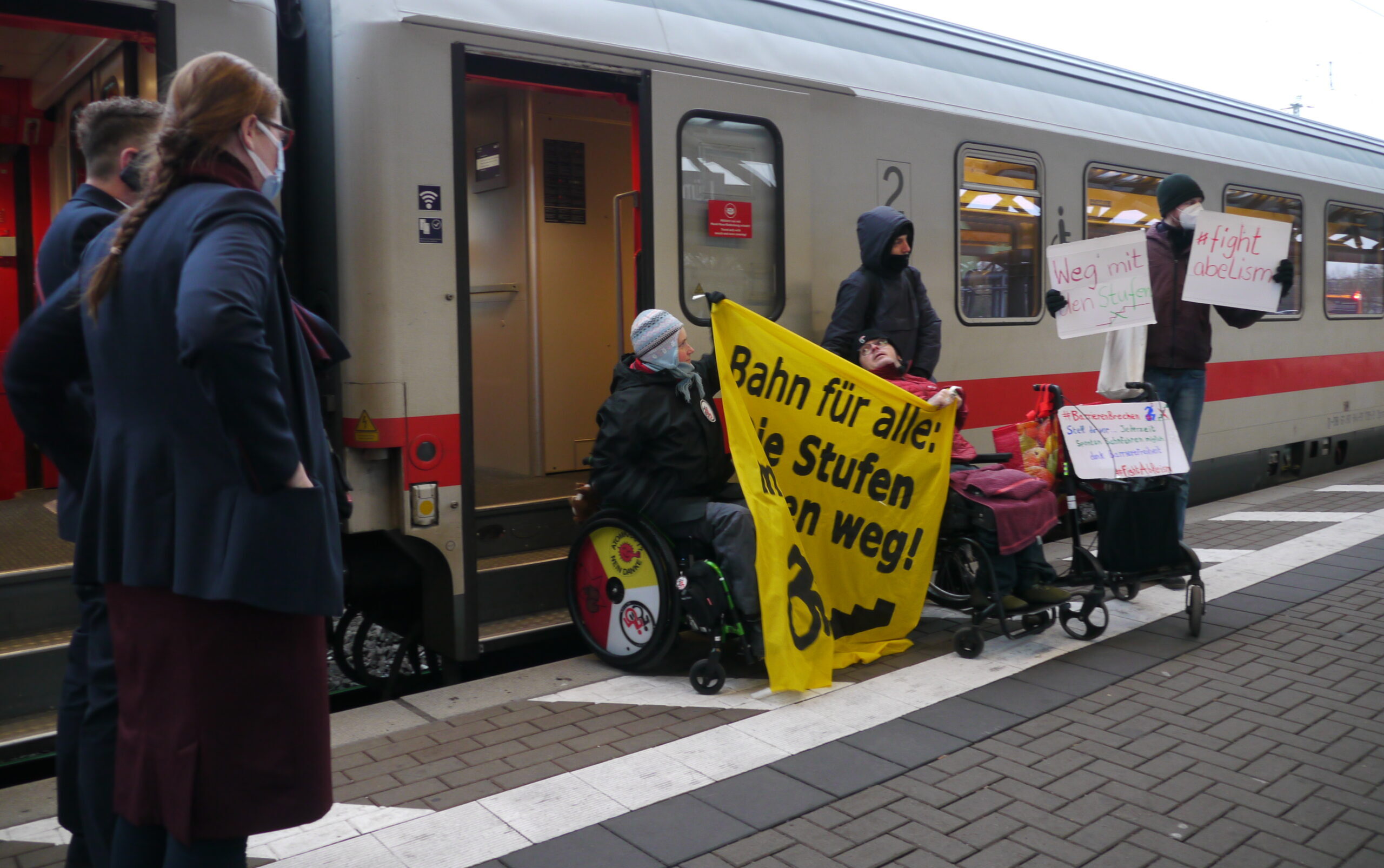 Protestaktion u.a mit Banner "die Stufen müssen weg" vor der Tür eines IC,, zwei Menschen im Rollstuhl dabei zwei Schaffner*innen gucken in die Richtung der Aktion
