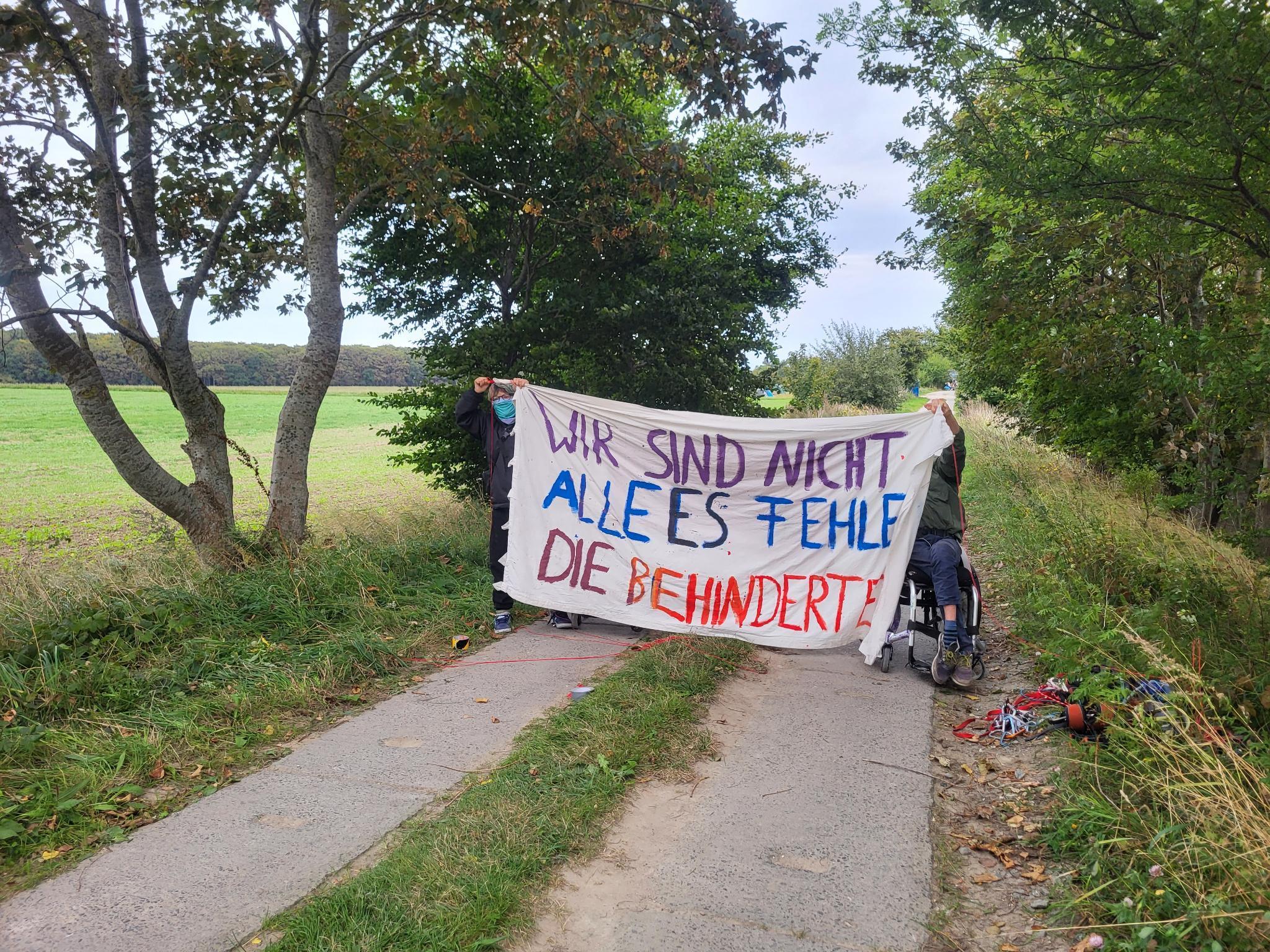 2 Menschen auf einem feld weg, eine davon im Rollstuhl halten ein Banner "wir sind nicht alle es fehlen die Gefangenen"