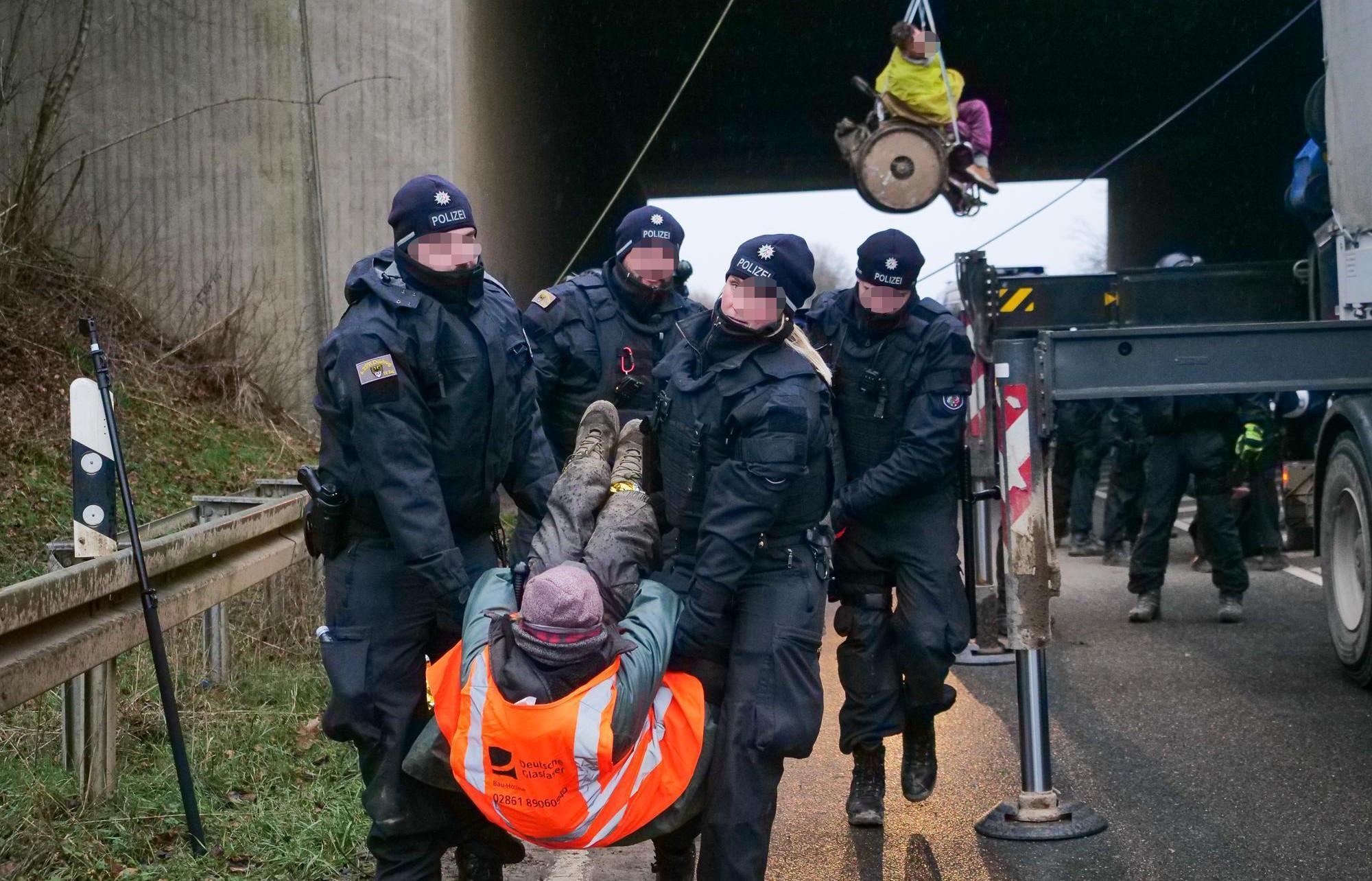 Lützerath, eine Person wird von der Polizei weg getragen. Im Hintergrund eine kletternde Person im Rollstuhl an einer Brücke im Seil