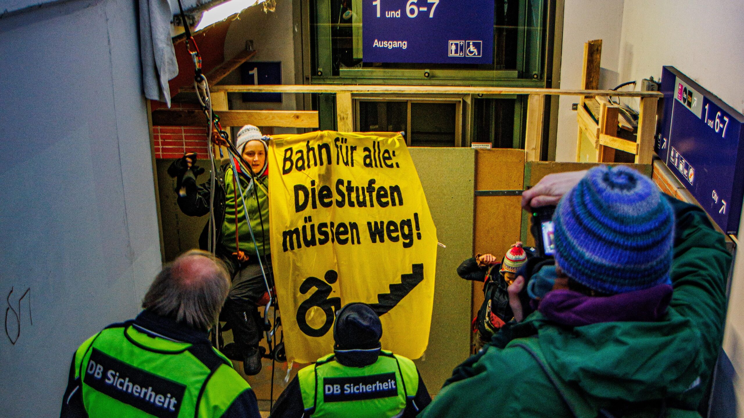 eine Person hängt im Rollstuhl sitzend in Seilen an einer Bahnunterführung, hinter ihr eine Aufzugbaustelle, davor die Treppe zum Gleis und DB Siherheit. Die Person im Seil hält ein Baner: die Stufen müssen weg mit Rollstuhl fahrender Person vor einer Treppe
