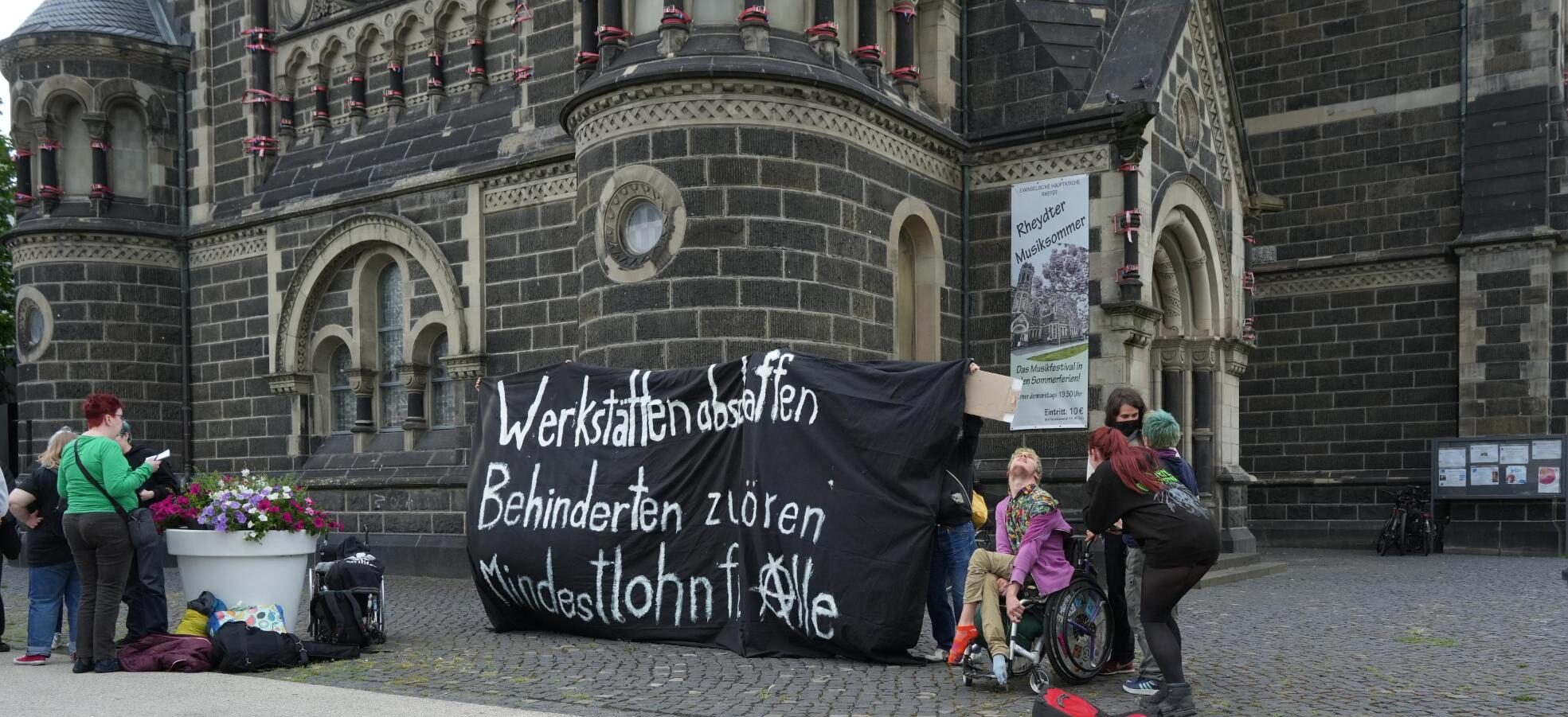 Menschen vor der Kirche mit einem Banner "Werkstätten abschaffen, Behinderten zuhören, Mindeslohn für Alle (mit Anarchie A). Links davon, Menschen die Infomaterial lesen.