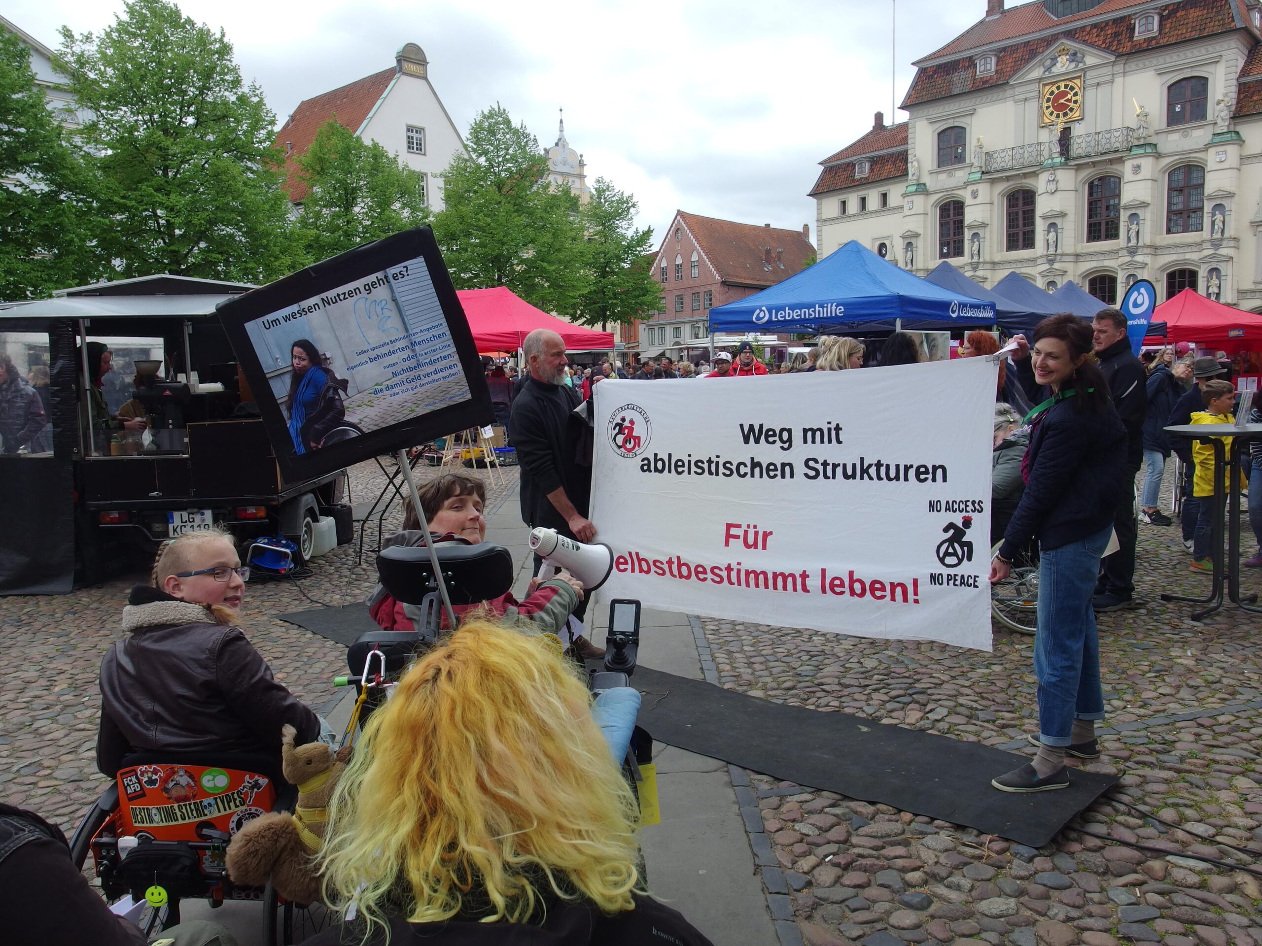 Menschen mit und ohne Rollstuhl mit Megafon, Banner "Weg mit ableitischen Strukturen. Für selbstbestimmt leben! im Hintergrund ein Stand der Lebenshilfe