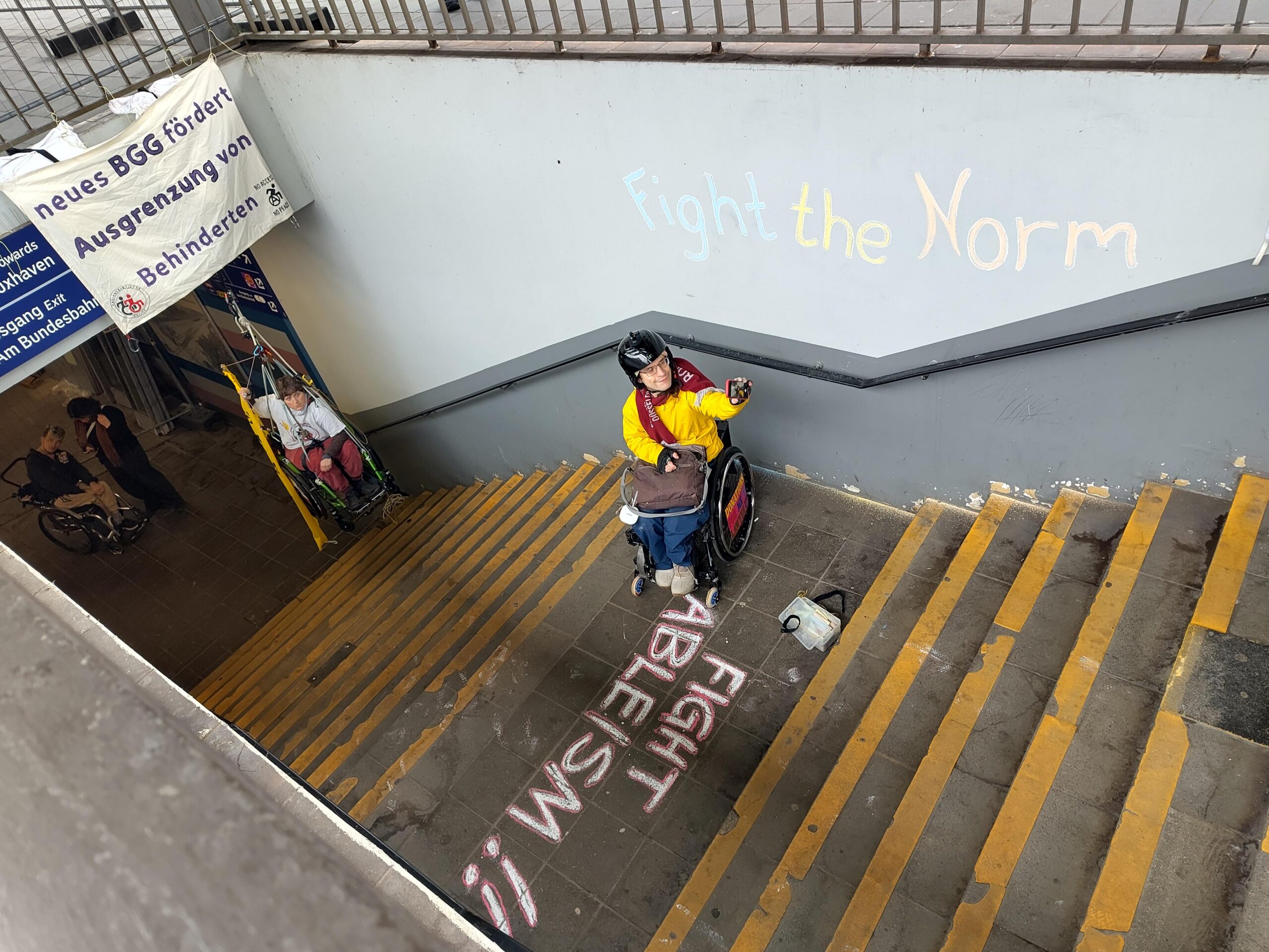 Blick auf das Treppenhaus am Bahnhof Buxtehude. Hintergrund, eine Person im Rollstuhl in der Luft im Seil gesichert, im Vordergrund 1 Mensch mit Rollstuhl. und Kreisde Spruch "Fight Ableism" auf der Treppe, an der Wand "Fight the Norm"