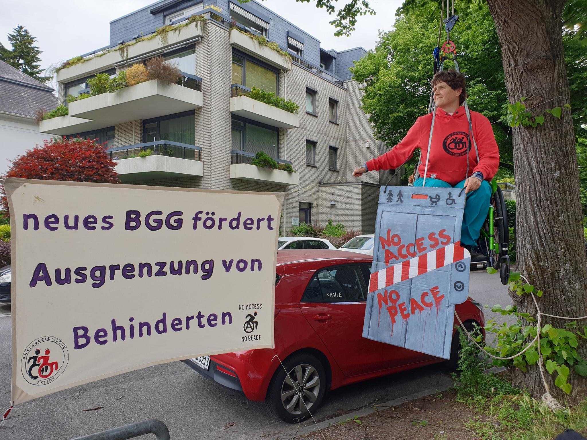 1 person sitz in seilen gesichert im Rollstuhl in der Ludt. Sie hält einen Aufzug aus Pappcarton in der Hand "no access no peace" daneben ein Banner: neues BGG fördert Ausgrenzung von Behinderten"