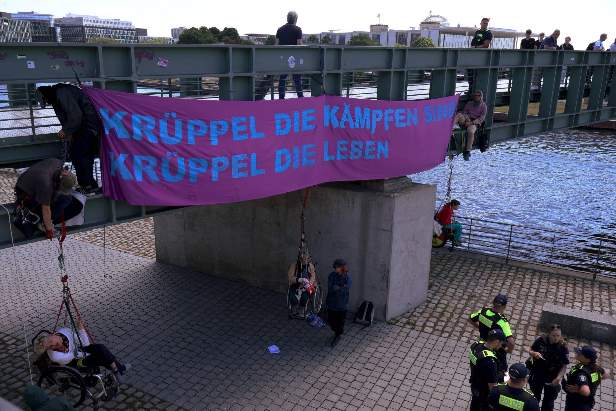 großes lila Banner "Krüppel die kämpfen sind Krüppel die leben" an einer Brücke 6 menschen klettern, 3 davon in seilen im rollstuhl sitzend gesichert. im Hintergrund ist der Reichtstag zu sehen
