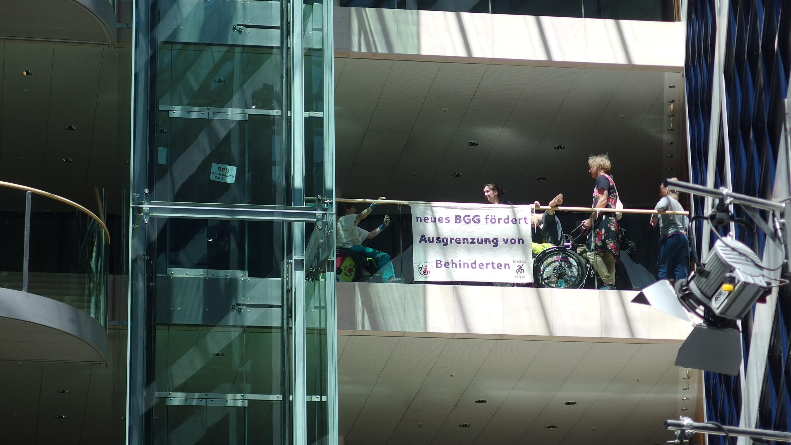 menschen sind an der Ballustrade aus Glas von einem Stockwerk zu sehen, einige haben einen Rollstuhl. Sie haben ein Banner an der Ballustrade angebracht "neues BGG fördert Ausgrenzung von Behinderten". und links beim Glasaufzug ein Schild "SPD mach keinen Scheiß"